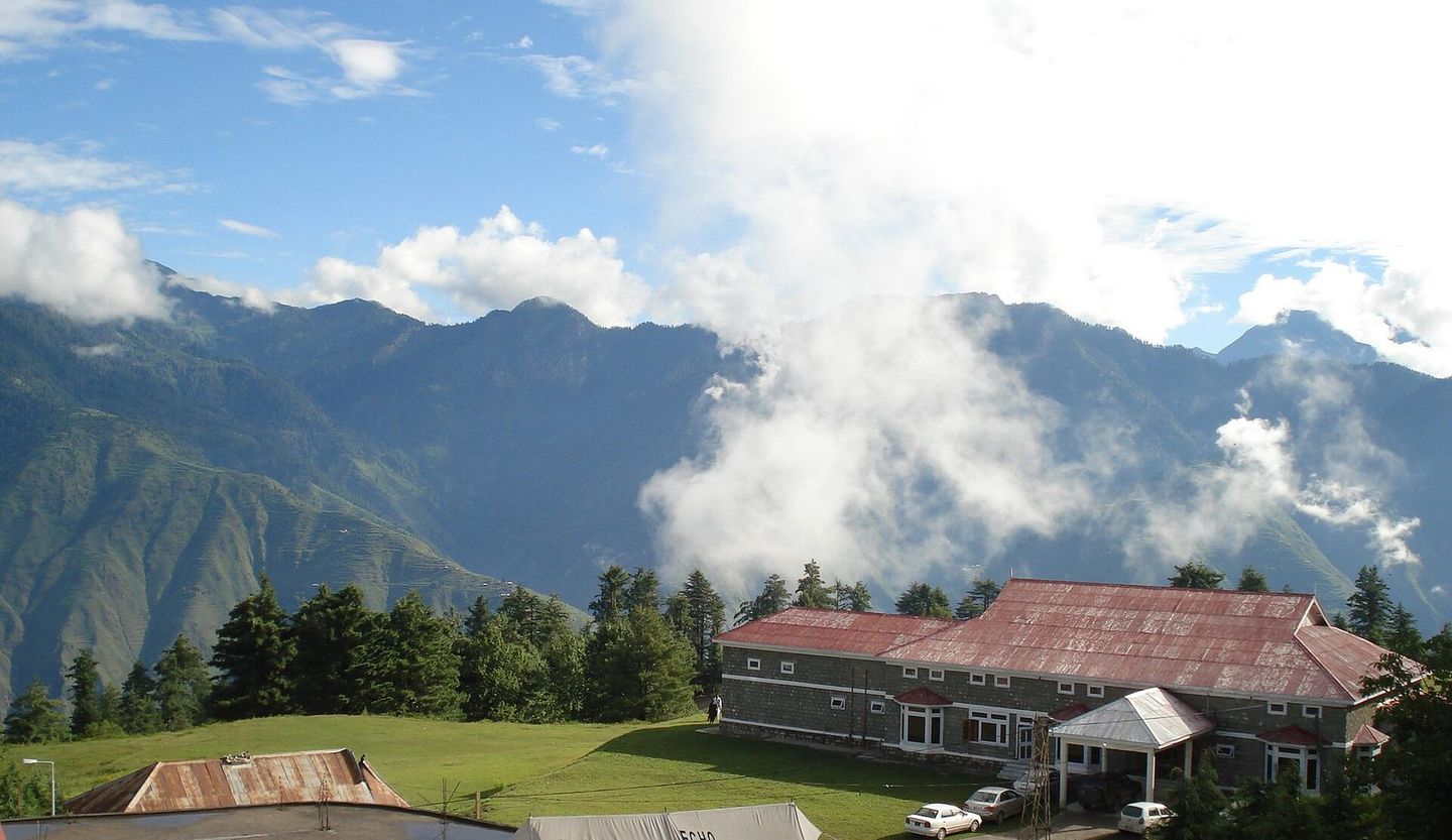 A red-roofed rest house among pines at Shogran, with clouds over the mountains