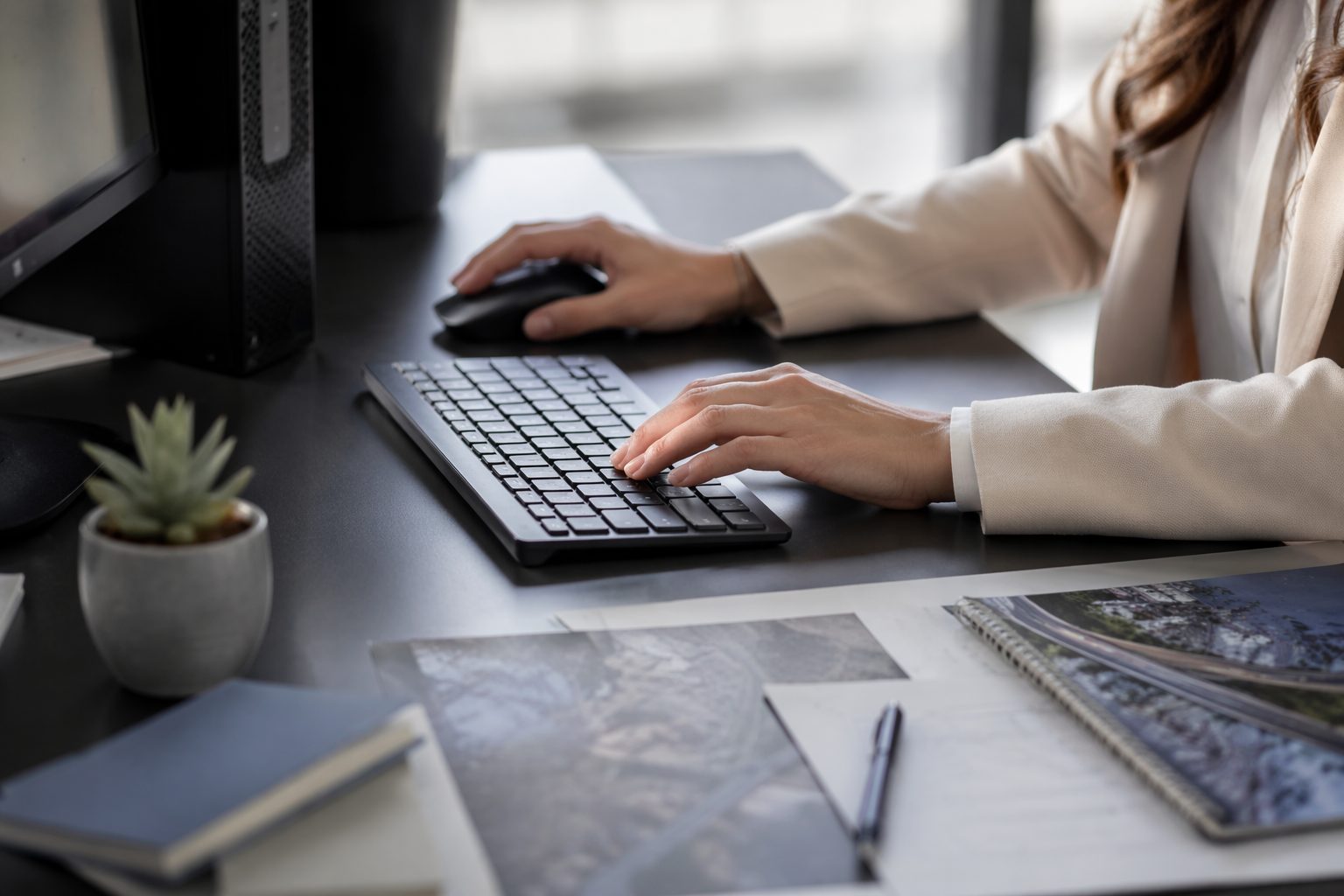 Close-up of a young woman’s hands on a mouse and keyboard beside a PC, with neatly arranged planning materials on a desk.