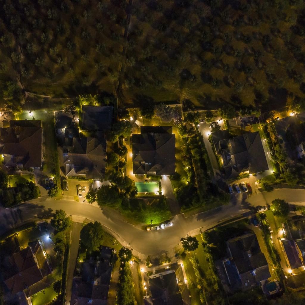 Fotografía aérea nocturna de un olivar en la Toscana con hileras ordenadas, luces urbanas al fondo y paleta tropical vibrante.