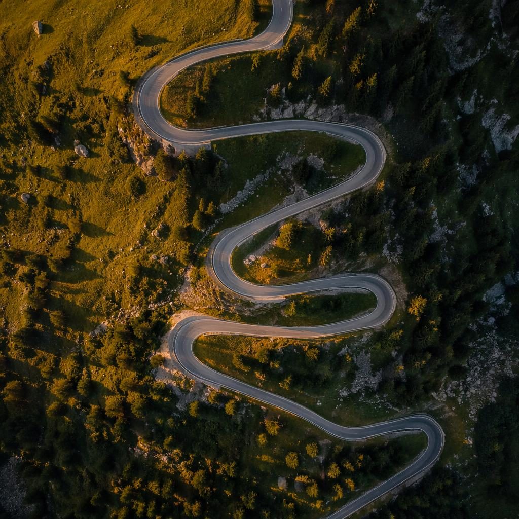 Top-down drone view of alpine highway switchbacks through golden-hour wine country with sharp, realistic detail.