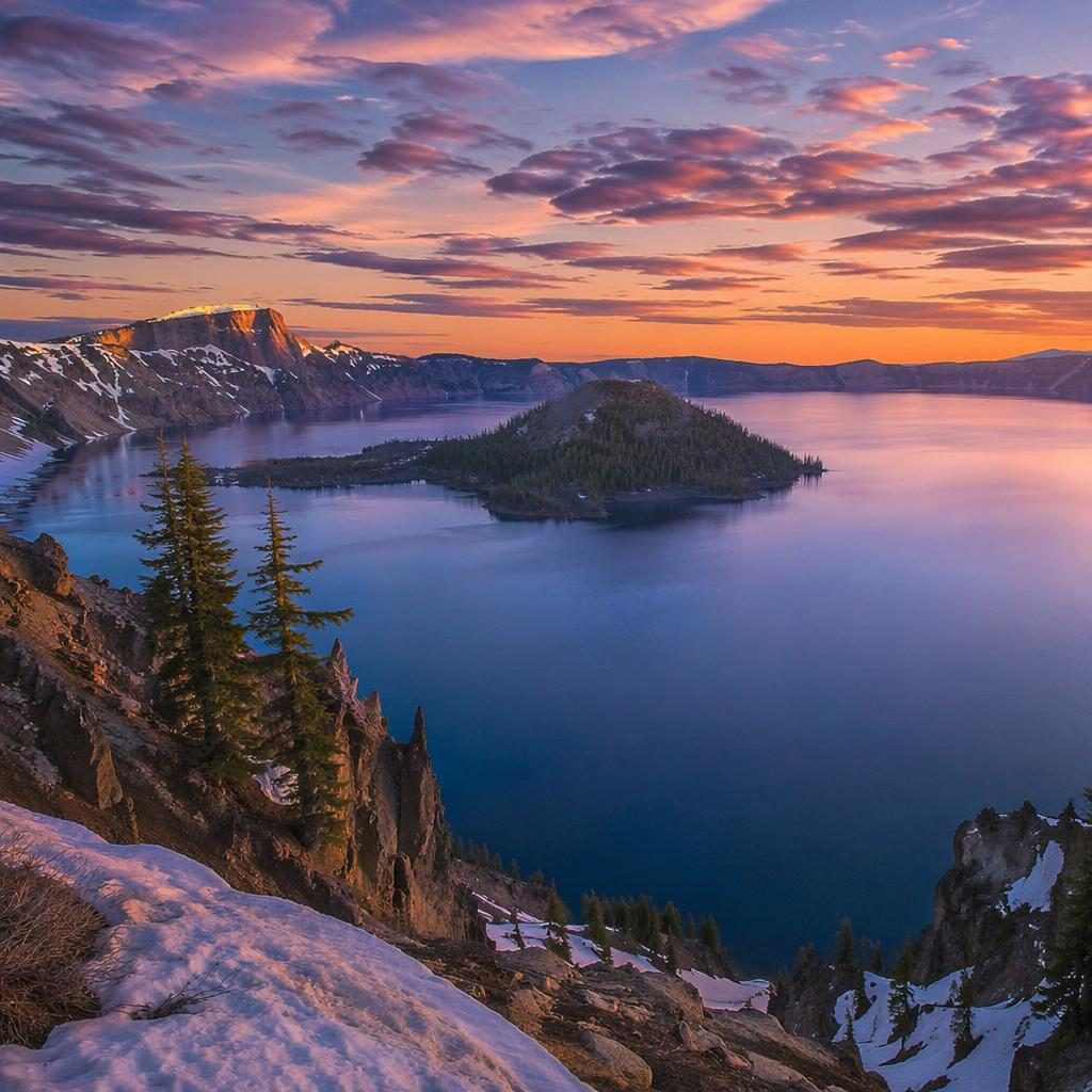 Blue hour landscape of a clear blue crater lake in Oregon, with volcanic slopes, spring snow, and warm twilight light.