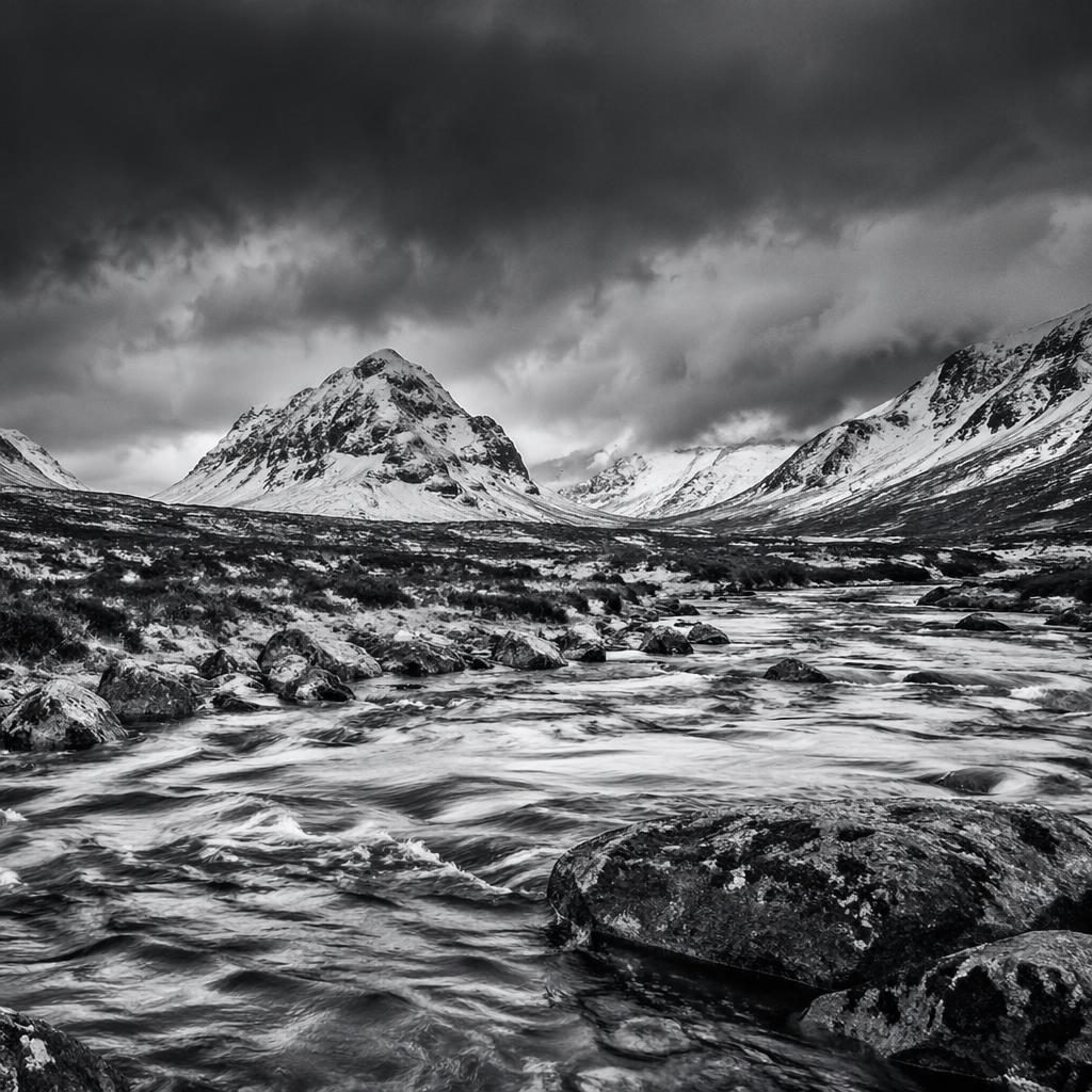 Black and white winter river valley in the stormy Scottish Highlands at dusk with dramatic clouds and sharp mountain ridges.