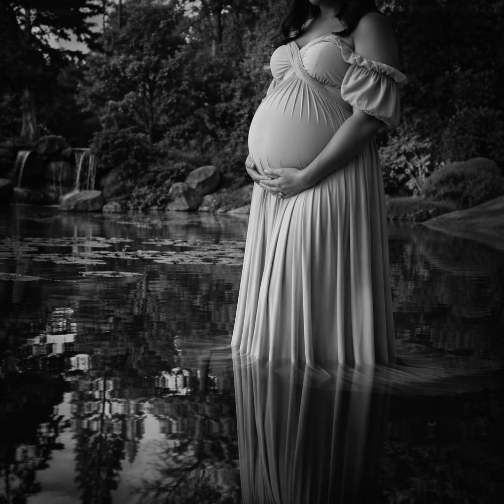 Close-up black and white maternity photo of a pregnant belly in a botanical garden beside a calm reflective lake