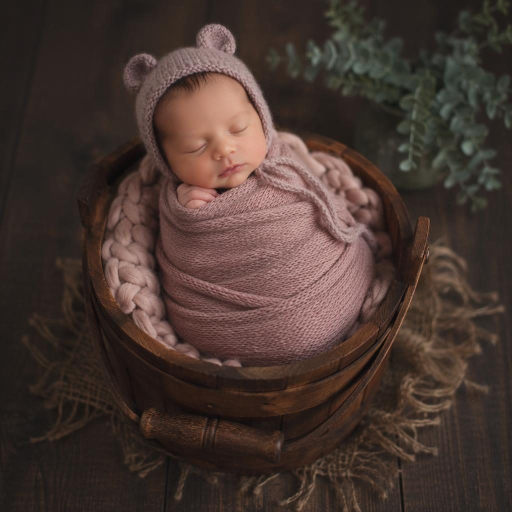 Wrapped newborn in a wooden basket for photoshoot, tiny knit hat, parent hands cradling, moody studio light