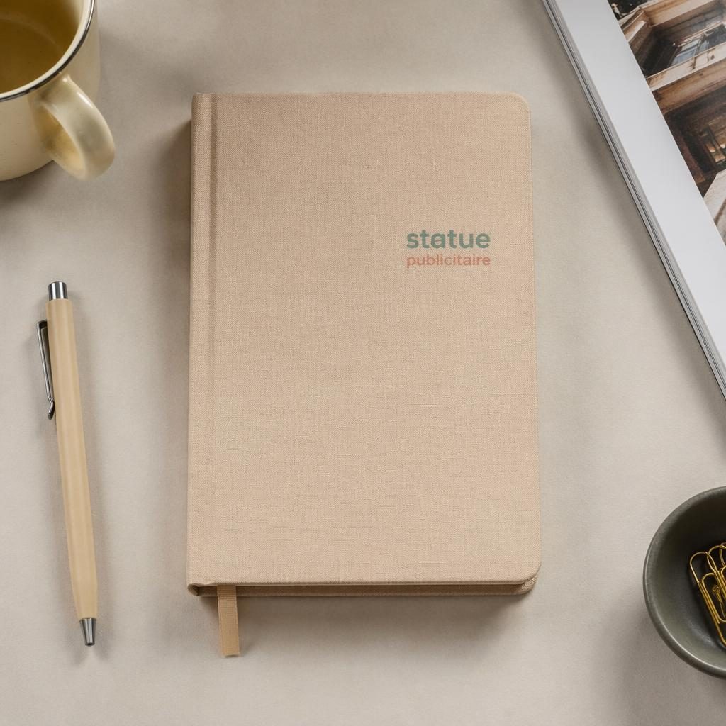 Carnet beige en mockup photo réaliste, vu à plat en studio, avec petit logo statue publicitaire et texture papier visible.