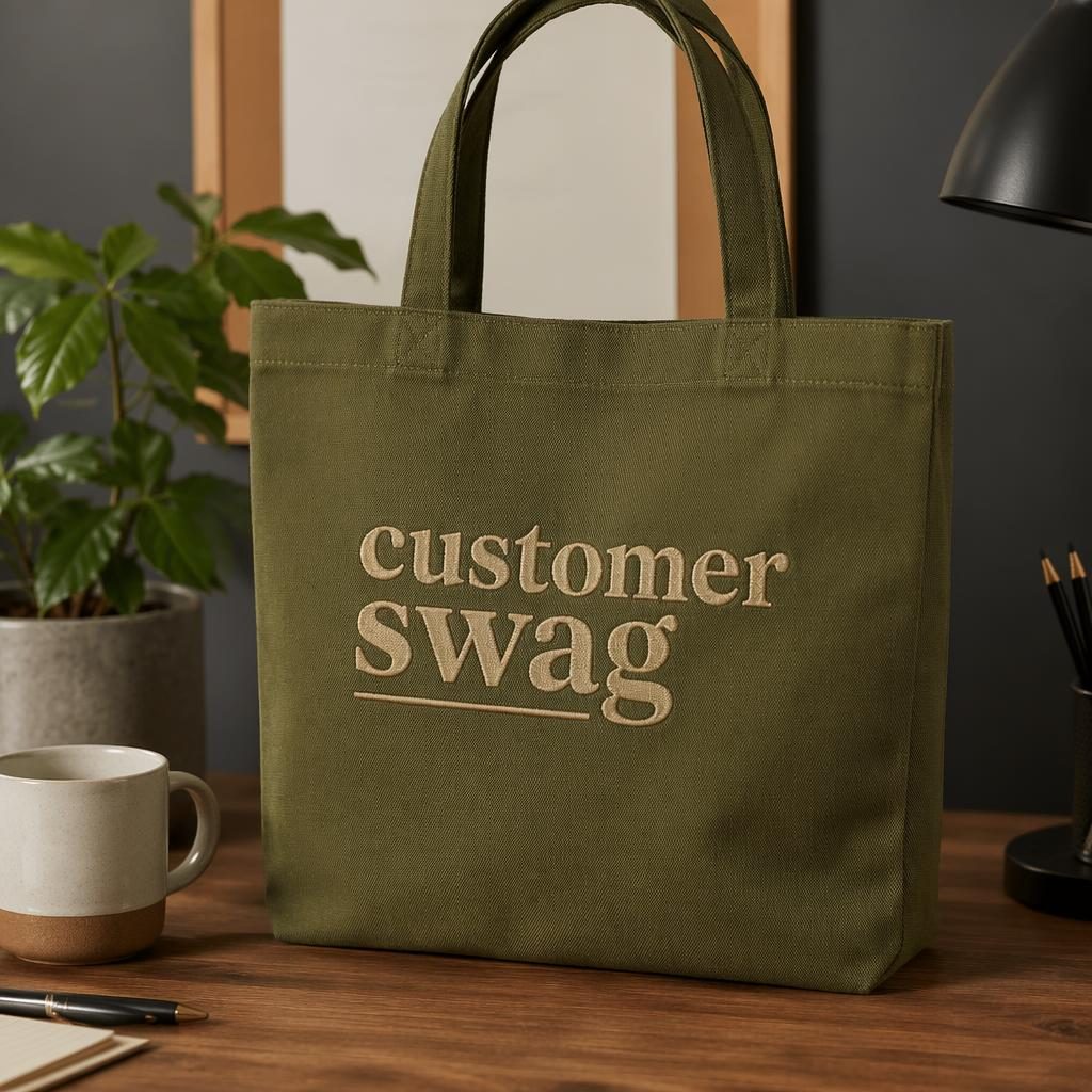 Olive green tote bag mockup with customer swag embroidery on a warm-toned desk lifestyle setup.