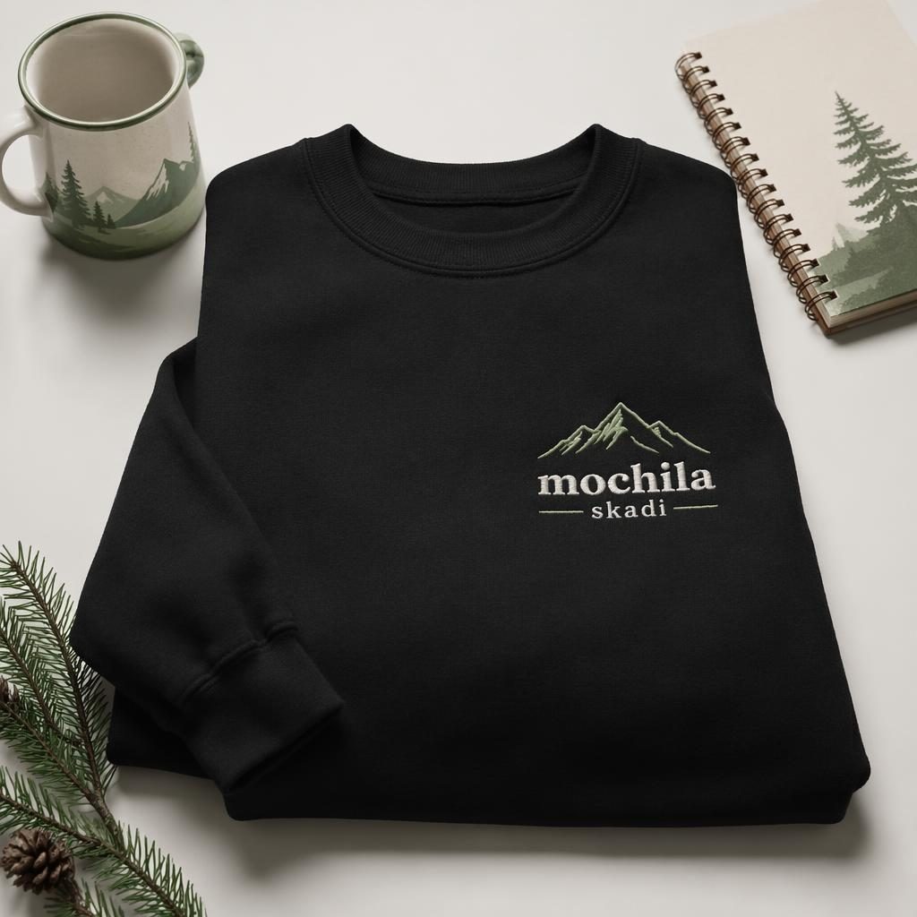 Sudadera negra en mockup fotográfico sobre fondo blanco, con logo bordado mochila skadi en tonos salvia y marfil.