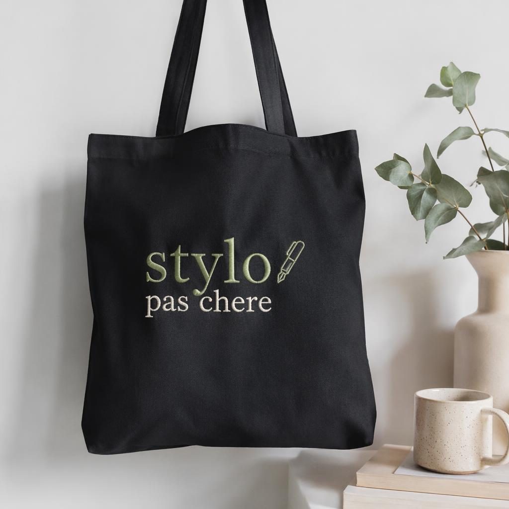 Tote bag noir en mockup photo réaliste sur fond blanc, logo brodé sage et ivoire, texture du tissu nette.