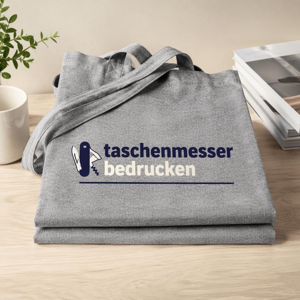 Gefalteter heather-grauer Tote Bag als Fotomockup mit vollflächigem Rückenlogo in Navy und Creme im Studio