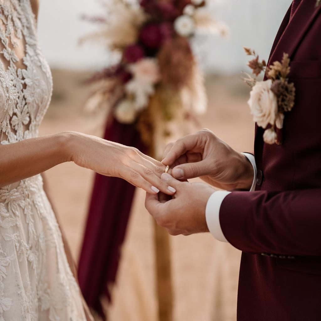 Nahaufnahme eines Ringtauschs bei einer Hochzeit mit Wunderkerzen in minimalistischer Wüstenkulisse, Pastelltönen, Bordeaux und Gold.