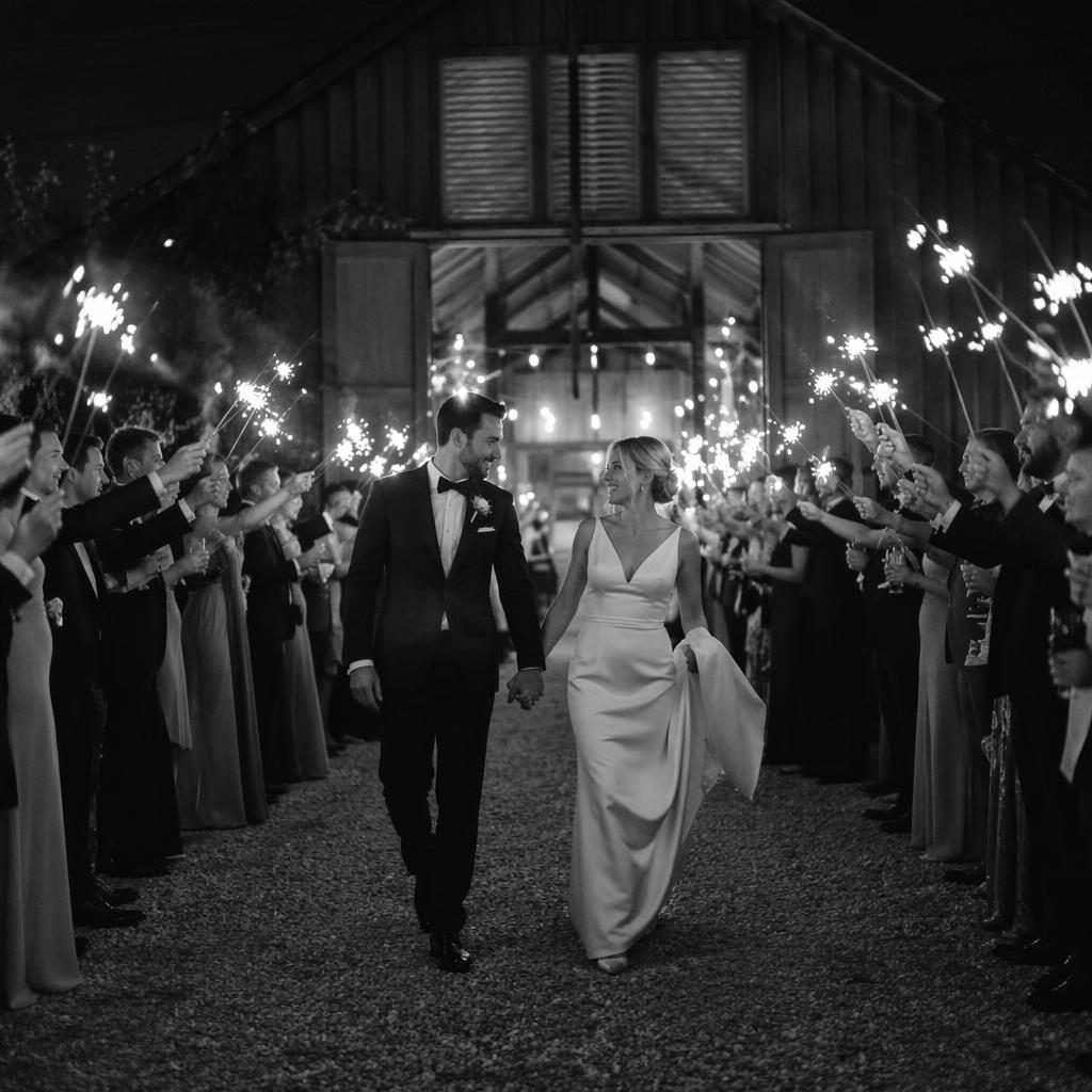 Black and white wedding photo of a couple walking through sparklers near a rustic barn, styled as an editorial elopement scene.