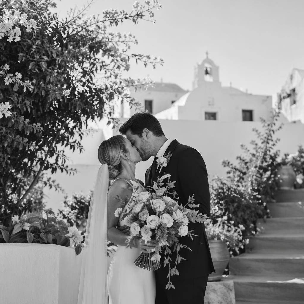 Couple de mariés s’embrassant dans un jardin botanique, lieu grec blanchi à la chaux, bouquet de mariée visible, noir et blanc.