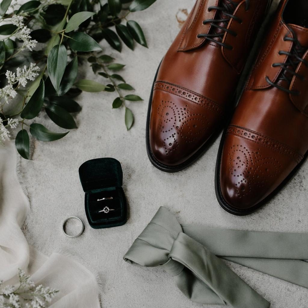Bodegón de zapatos del novio y anillos de boda sobre fondo blanco y verde salvia en jardín botánico con luz natural.