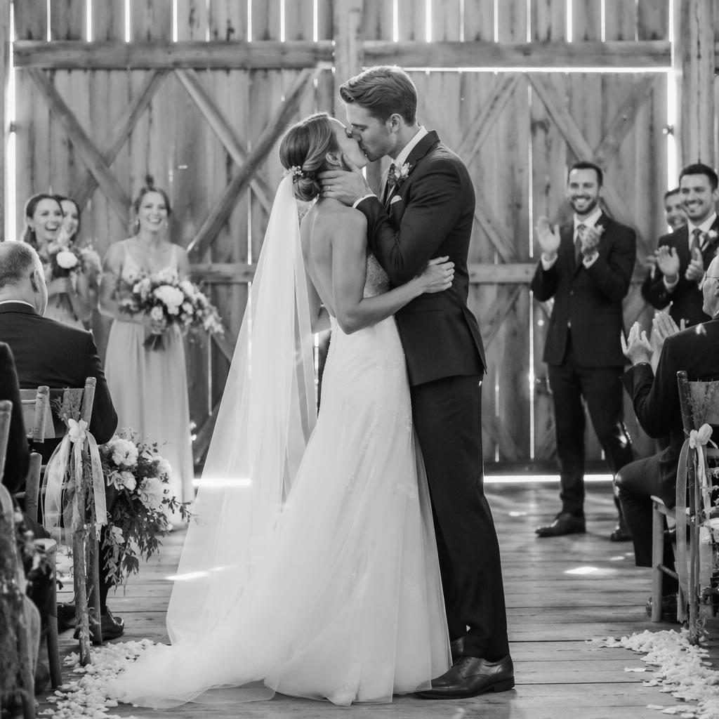 Black and white wedding photo of a couple sharing a first kiss at a rustic barn with natural light and editorial framing.