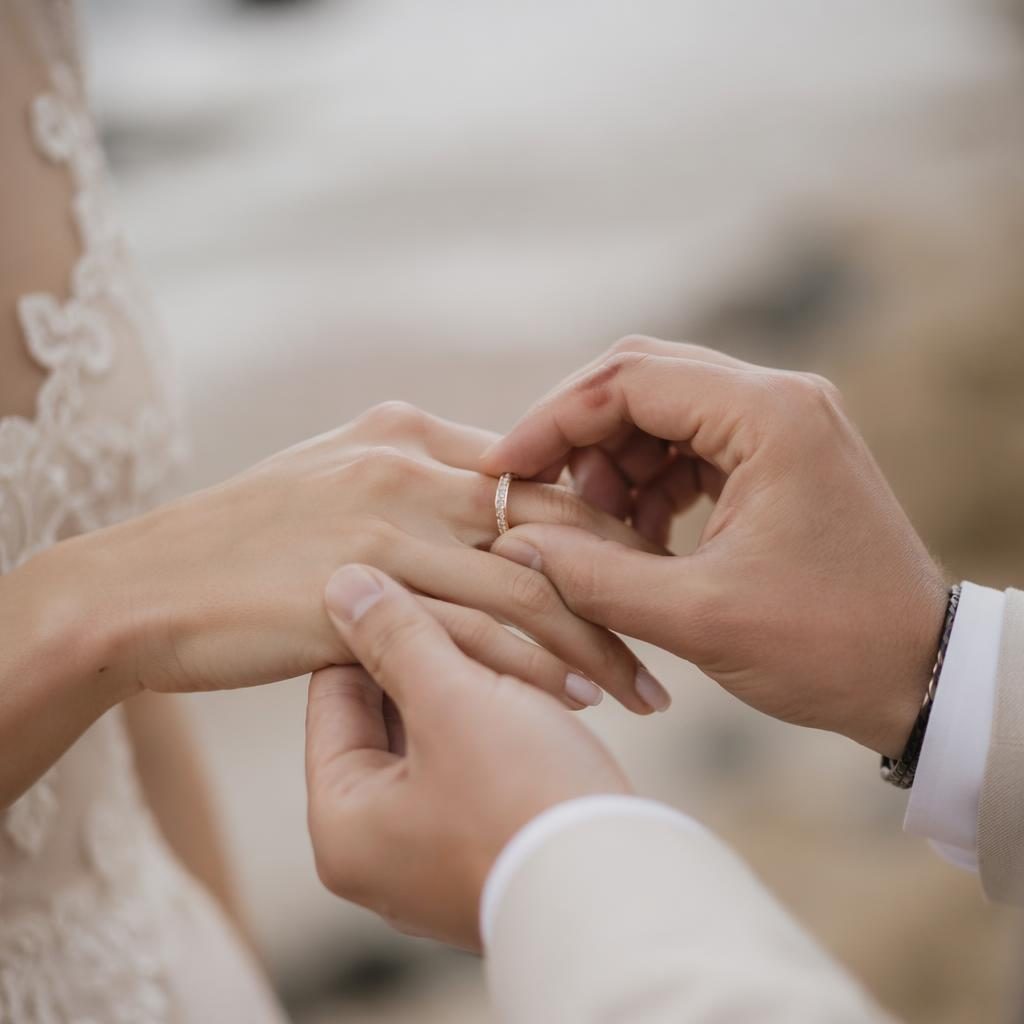 Primer plano de manos intercambiando anillos en una boda en la playa, con luz natural y tonos burdeos y dorado.