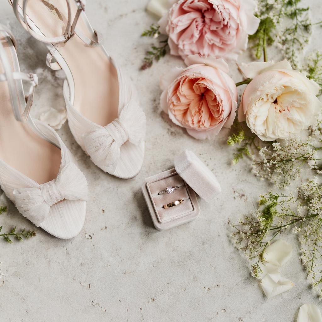 Bodegón de boda con zapatos y anillos sobre detalles de recepción frente al acantilado y el mar, en tonos blush y crema.